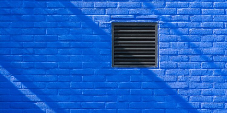 Close-up of a bright blue brick wall featuring a black ventilation grate and shadows.