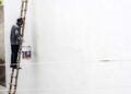 A worker on a ladder paints a large white wall outdoors, showcasing construction work.