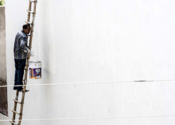 A worker on a ladder paints a large white wall outdoors, showcasing construction work.