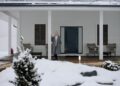 Elderly woman in coat standing on a snowy porch during winter, surrounded by snow-covered landscape.