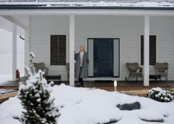 Elderly woman in coat standing on a snowy porch during winter, surrounded by snow-covered landscape.