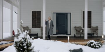 Elderly woman in coat standing on a snowy porch during winter, surrounded by snow-covered landscape.