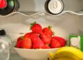 A bowl of ripe strawberries in a refrigerator, surrounded by drinks and bananas, showcasing vibrant freshness.