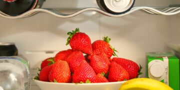 A bowl of ripe strawberries in a refrigerator, surrounded by drinks and bananas, showcasing vibrant freshness.