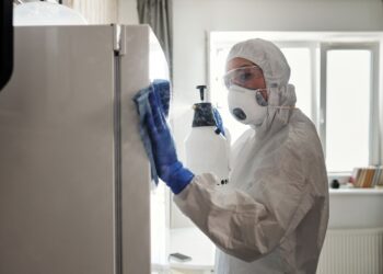 Person in protective gear sanitizing refrigerator at home for safety.