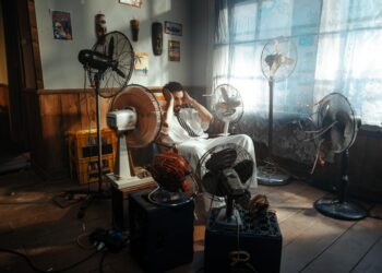 Man in white outfit sitting surrounded by several fans indoors during daytime.