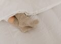 Comfortable feet in woolen socks peek out under a soft white linen blanket on a bed.