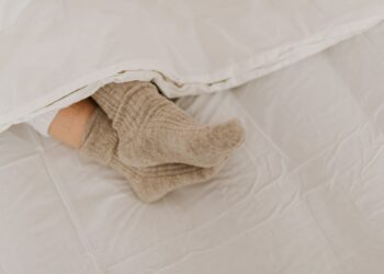 Comfortable feet in woolen socks peek out under a soft white linen blanket on a bed.