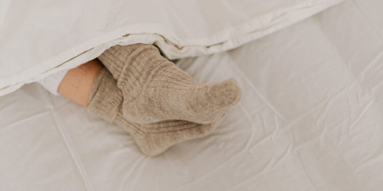 Comfortable feet in woolen socks peek out under a soft white linen blanket on a bed.