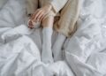 An adult enjoying a warm drink while relaxing in a cozy bed with a knitted blanket and white socks.