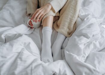 An adult enjoying a warm drink while relaxing in a cozy bed with a knitted blanket and white socks.