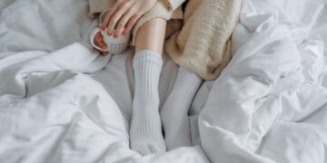 An adult enjoying a warm drink while relaxing in a cozy bed with a knitted blanket and white socks.