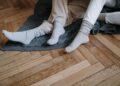 A couple wearing socks, relaxing on a wooden floor with a gray blanket, enjoying warmth.