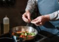 Chef seasoning a vegetable stir fry in a pan with fresh herbs on a stove.