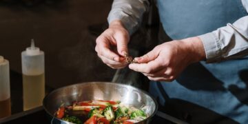 Chef seasoning a vegetable stir fry in a pan with fresh herbs on a stove.