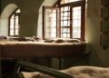 Sunlight streams through open windows onto a cozy bed in a rustic room, creating a warm atmosphere.