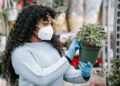 Side view of African American female in protective mask and gloves with green potted plant in hands in floral market with various flowers on blurred background
