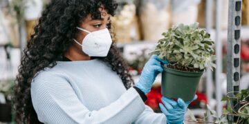 Side view of African American female in protective mask and gloves with green potted plant in hands in floral market with various flowers on blurred background