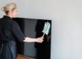 A female housekeeper performing dusting on a television screen indoors.