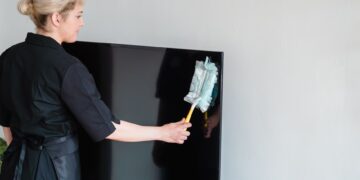 A female housekeeper performing dusting on a television screen indoors.